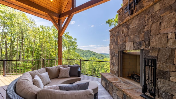 A covered outdoor seating area with a stone fireplace, surrounded by a wooden deck and overlooking a forested landscape.