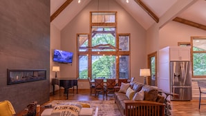 A spacious living room with a high ceiling, wooden beams, a large window with a view of trees, a piano, a sofa, and a dining area.