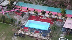 A rectangular pool with a blue interior, surrounded by a tiled deck. There are multiple buildings with colorful roofs in the background. - Camp Awara Rishikesh (Narendranagar)