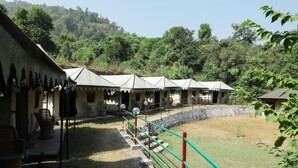 A row of simple, single-story buildings with a flat roof, surrounded by a grassy area and trees. - Camp Awara Rishikesh (Narendranagar)