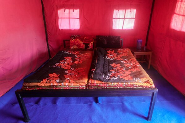 A tent-style room with two beds, each with floral bedding. There are two windows with curtains, and a small table with a lamp.