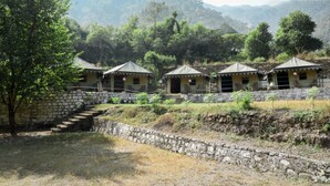 A row of rustic, single-story cabins with stone walls and thatched roofs, set against a backdrop of lush greenery and hills. - Camp Awara Rishikesh (Narendranagar)