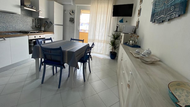 A modern kitchen with white cabinets, a dining table with blue chairs, a potted plant, and a flat-screen TV mounted on the wall. - Apartment 'Maison Admas' with Mountain View, Balcony and Air Conditioning (Villasimius)
