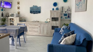 A living room with a blue sofa, a dining area with a table and chairs, a kitchen area with a microwave, and a TV mounted on the wall. - Apartment 'Maison Admas' with Mountain View, Balcony and Air Conditioning (Villasimius)