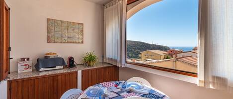 Un coin repas avec une table dressée pour deux personnes, une vue sur la mer, une carte accrochée au mur et un plan de travail de cuisine avec un grille-pain et une plante.