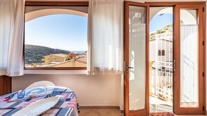 A bedroom with a bed, a window with curtains, a door leading to a balcony, and a view of a building and hills. - Apartment 'Adriana's House' with Sea View, Private Terrace and Air Conditioning (Buggerru)