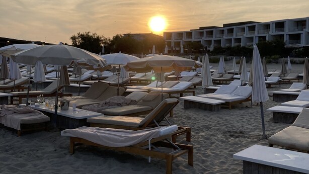 En strand med solstolar och parasoller, en byggnad i bakgrunden och solen som går ner.