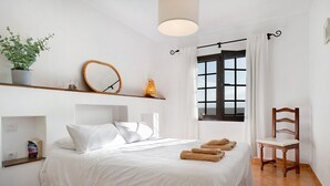 Un lit bien fait, avec du linge de lit blanc, une chaise en bois, une étagère avec une plante, un miroir et une fenêtre avec des rideaux.