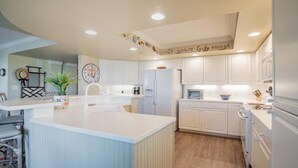A modern kitchen with white cabinetry, a central island, and a ceiling with a decorative banner. - Ocean Oasis Fordham 101 3BR Villa Ocean Views (Pawleys Island)
