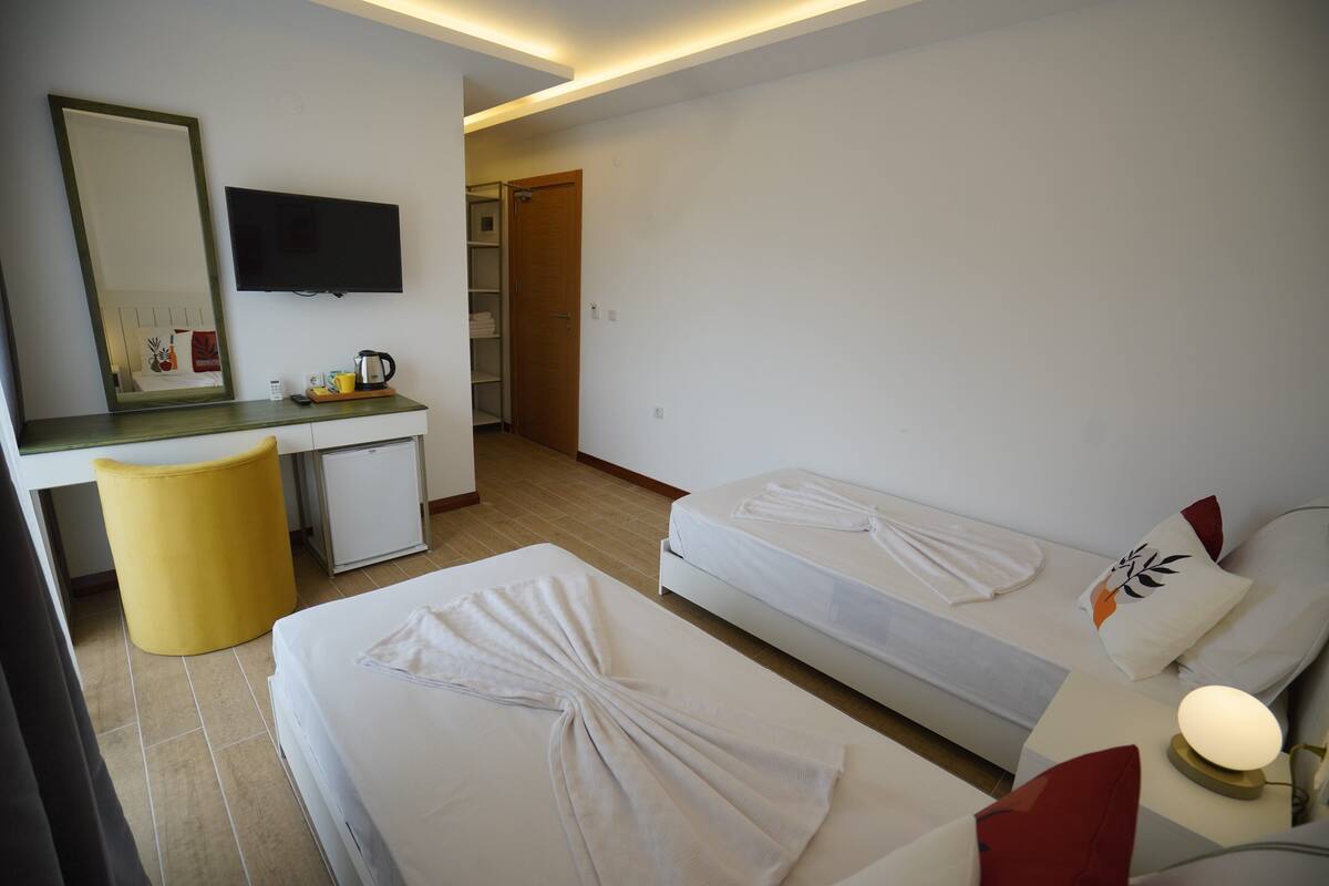 Une chambre d’hôtel avec deux lits simples, un bureau avec une chaise, une petite kitchenette et un téléviseur fixé au mur.