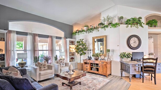 A living room with a grey sofa, a wooden coffee table, and a desk with a computer. There are plants on the wall and a large mirror above the desk. - Pool, Putting Green, Fire Pit: Elegant Peoria Home (Peoria)