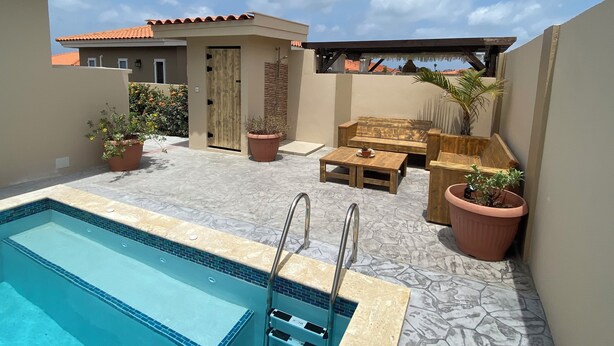 Ein Poolbereich mit Holzterrasse, einer Sitzecke mit Sofa und Couchtisch sowie ein kleines Gebäude mit einer Tür.