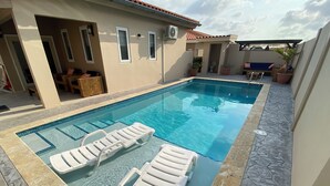 Ein Swimmingpool mit zwei weißen Liegestühlen, einem überdachten Sitzbereich und Topfpflanzen.