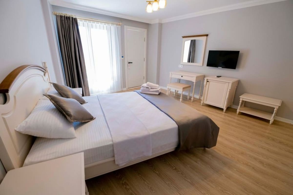 A hotel room with a bed, bedside tables, a desk, a chair, a television, and a window with curtains.