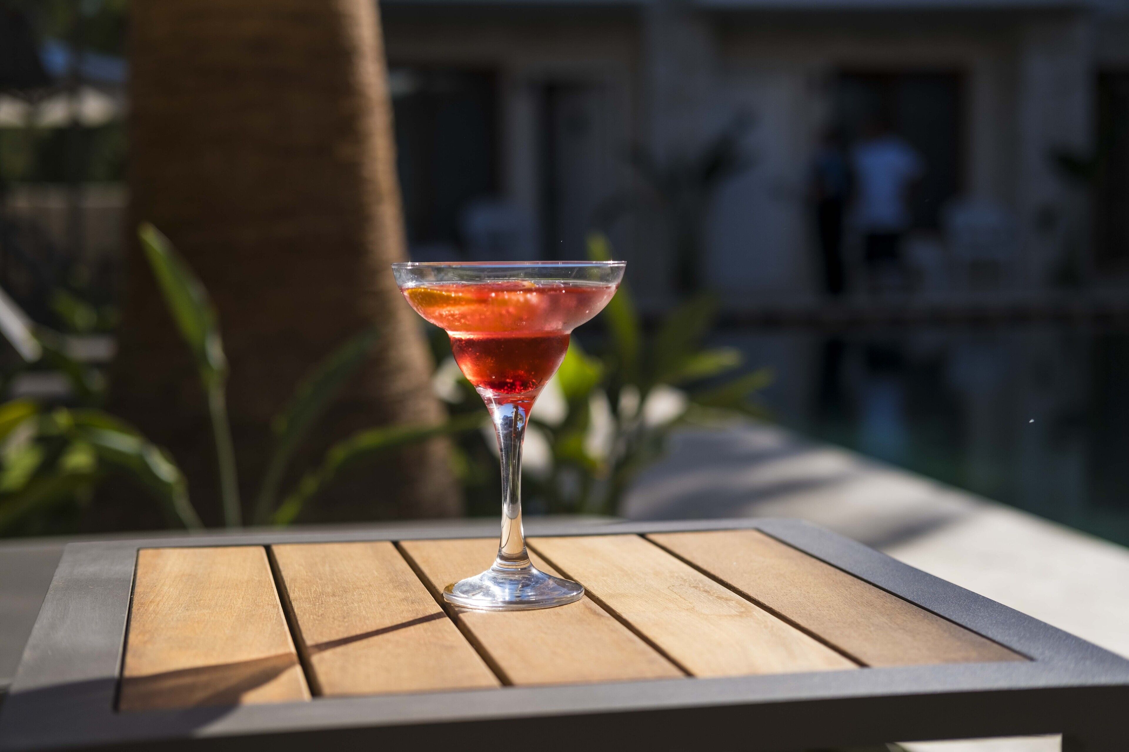 A cocktail glass on a wooden table with a blurred background of a building and greenery.