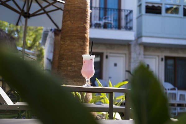 A glass of pink drink on a table with a palm tree and a building in the background. - ICARUS ALACATI (Cesme)