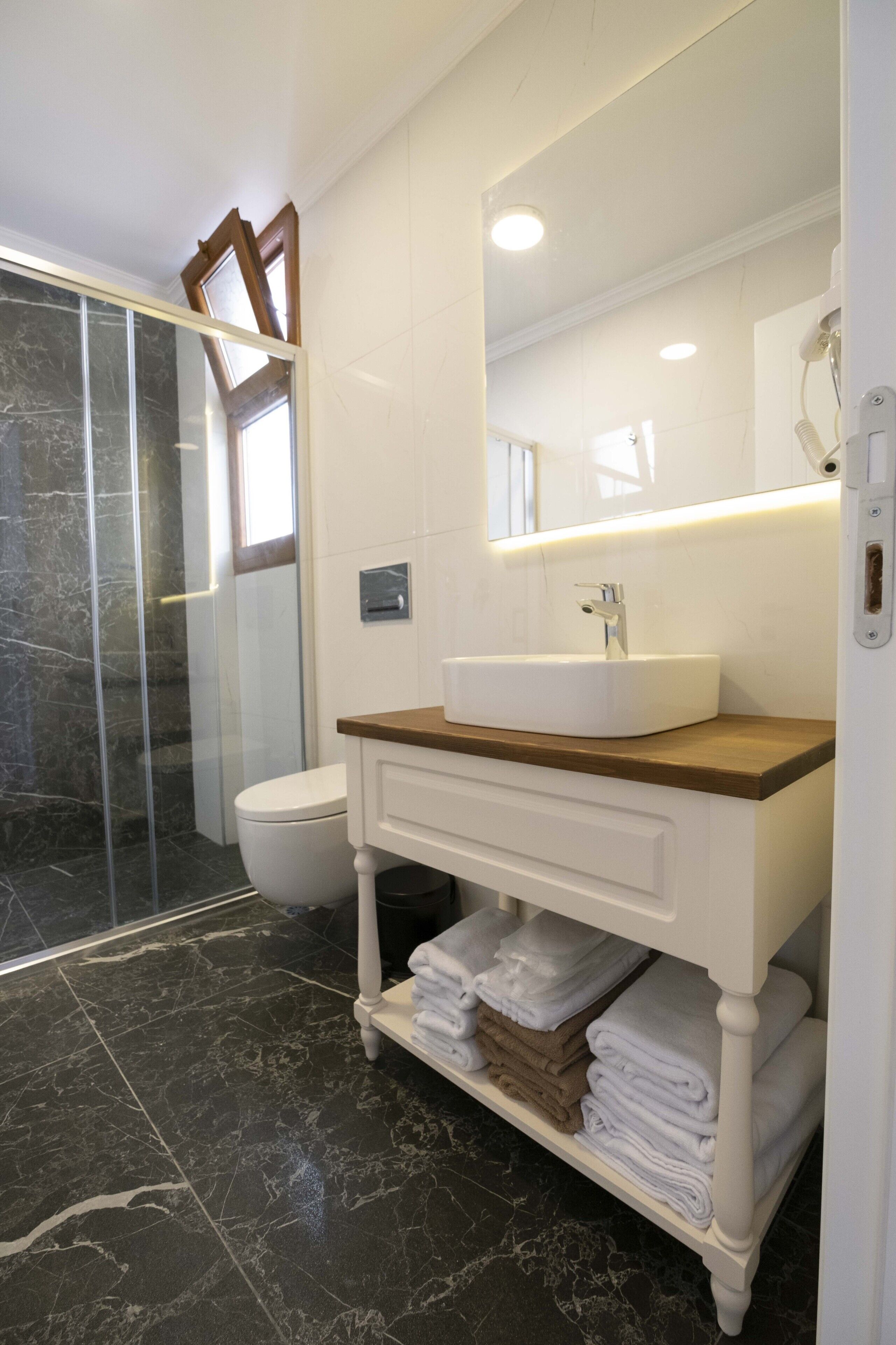 A bathroom with a marble floor, a white sink, a wooden vanity, a glass shower enclosure, and a toilet.