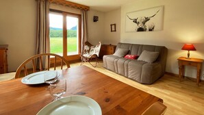 A dining area with a wooden table, chairs, and a sofa. There is a window with curtains, a framed picture on the wall, and a lamp on a side table. - Studio with mountain views and pools, pets allowed (Samoëns)
