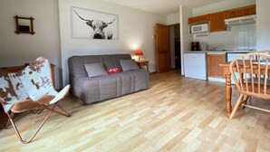 A living room with a grey sofa, a wooden chair, and a red cushion. A kitchen is visible in the background with wooden cabinets and a microwave. - Studio with mountain views and pools, pets allowed (Samoëns)