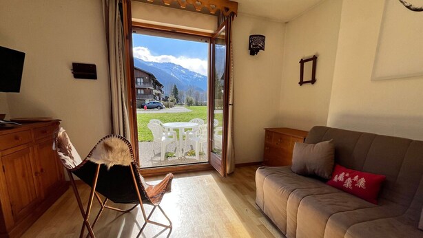 A living room with a sofa, a chair, a wooden cabinet, and a sliding glass door leading to an outdoor area with a table and chairs. - Studio with mountain views and pools, pets allowed (Samoëns)