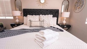 A neatly made bed with a quilted white bedspread, two black and white patterned pillows, and a folded white towel. There are two bedside tables with lamps, a large mirror above the bed, and decorative wall art. - New Pool/Spa & Pool Table @ 6BR Refined Vegas Stay (North Las Vegas)