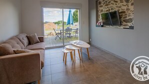 A modern living room with a sofa, a dining table with chairs, a TV mounted on a stone wall, and a sliding glass door leading to an outdoor patio.