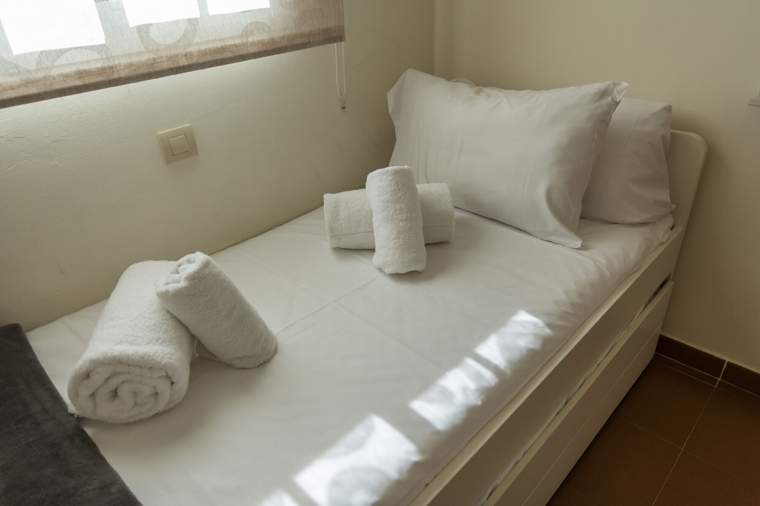 Een enkel bed met witte lakens, twee opgerolde handdoeken en een raam met gordijn.