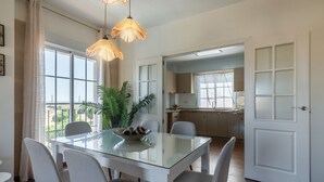 Un coin repas avec une table en verre, six chaises et un saladier rempli de fruits. On y trouve deux luminaires suspendus, une plante en pot et, par une porte ouverte, on aperçoit la cuisine.
