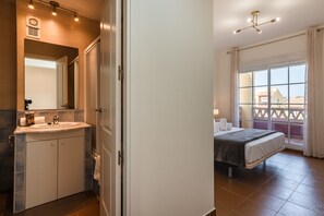 A hotel room with a bathroom featuring a sink, mirror, and towel rack. The room has a bed, a window with curtains, and a view of buildings outside. - Albatros Golf I - Costa Esuri EAHGI173AP111 (Ayamonte)