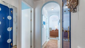 Un pasillo con una puerta azul, una puerta blanca y un arco azulado que conduce a una sala de estar con un sofá y una lámpara.