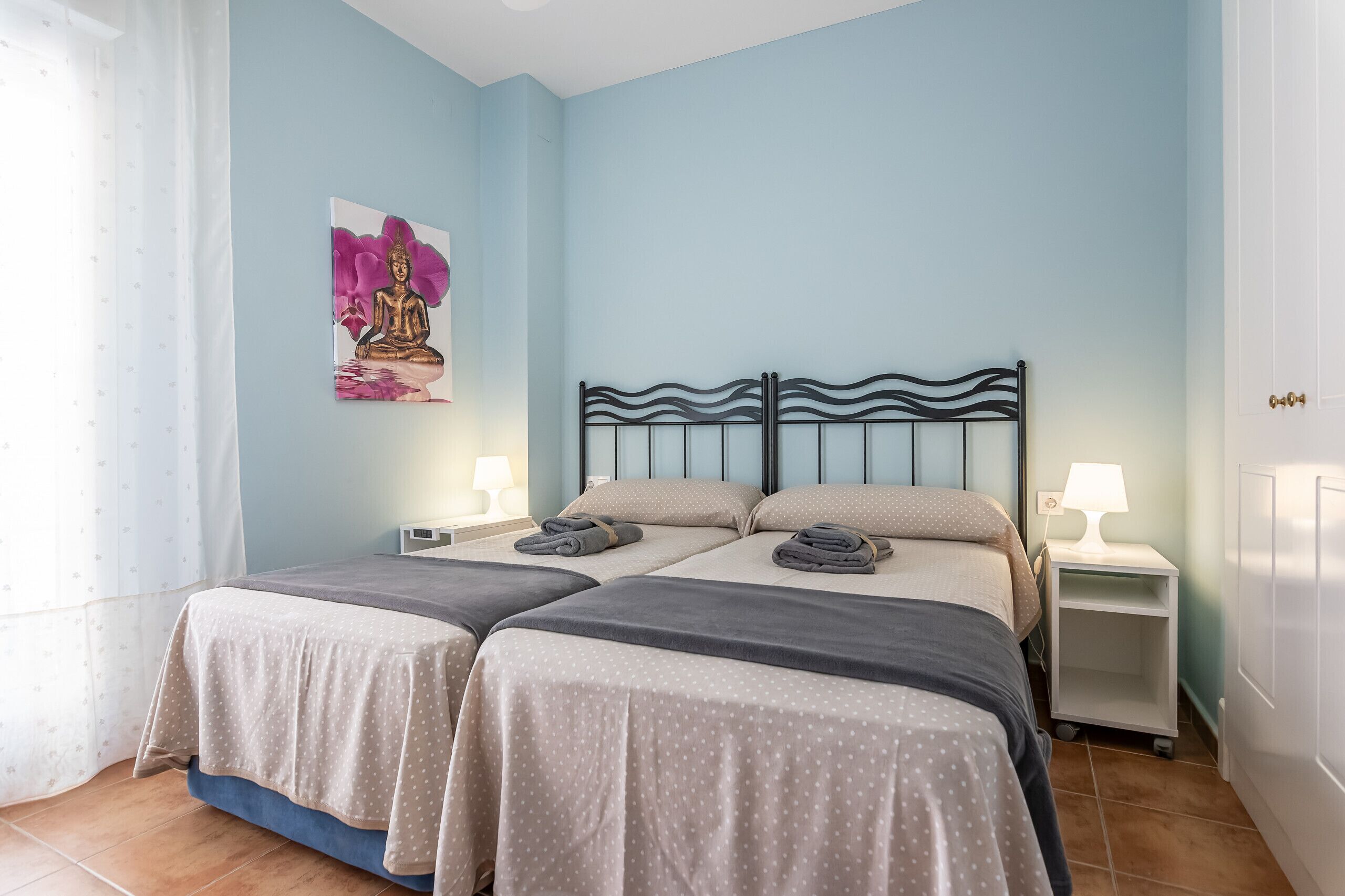 A double room with two beds, each with a pillow and a folded towel. There is a nightstand with a lamp on each side of the beds.