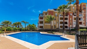 Una piscina rodeada de una terraza de ladrillo, con palmeras y un edificio de varios pisos al fondo.