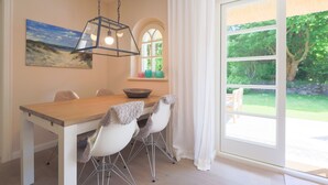 A dining area with a wooden table, white chairs, a hanging light fixture, a painting on the wall, and a view of a garden through a large window. - Reethüs - Keitum (Keitum)