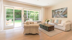 A spacious living room with a large sliding glass door leading to a garden, a striped sofa, a coffee table, and a painting on the wall. - Reethüs - Keitum (Keitum)