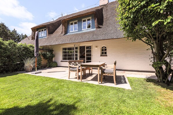 A house with a thatched roof, a covered patio with a table and chairs, and a well-maintained lawn.