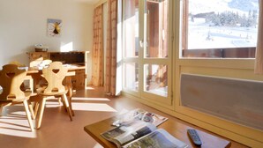 A room with a wooden desk, chairs, a table with magazines, a remote control, and large windows with a view of snow-covered mountains. - Divisible studio 4 pers, ski-in/ski-out, pets allowed (Plagne Bellecôte)
