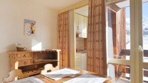 A dining area with a wooden table set for four, a wooden cabinet with drawers, a painting on the wall, and a view of a snowy landscape through a window. - Divisible studio 4 pers, ski-in/ski-out, pets allowed (Plagne Bellecôte)