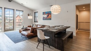 A modern living room with a wooden dining table, a brown leather sofa, and a white chair. - Eagle Lodge Shuttle | Mountain Views (Mammoth Lakes)