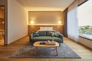 Habitación de hotel moderna con piso de madera, un sofá verde con cojines blancos, una mesa de centro de madera con libros y un florero, y un ventanal con cortinas.