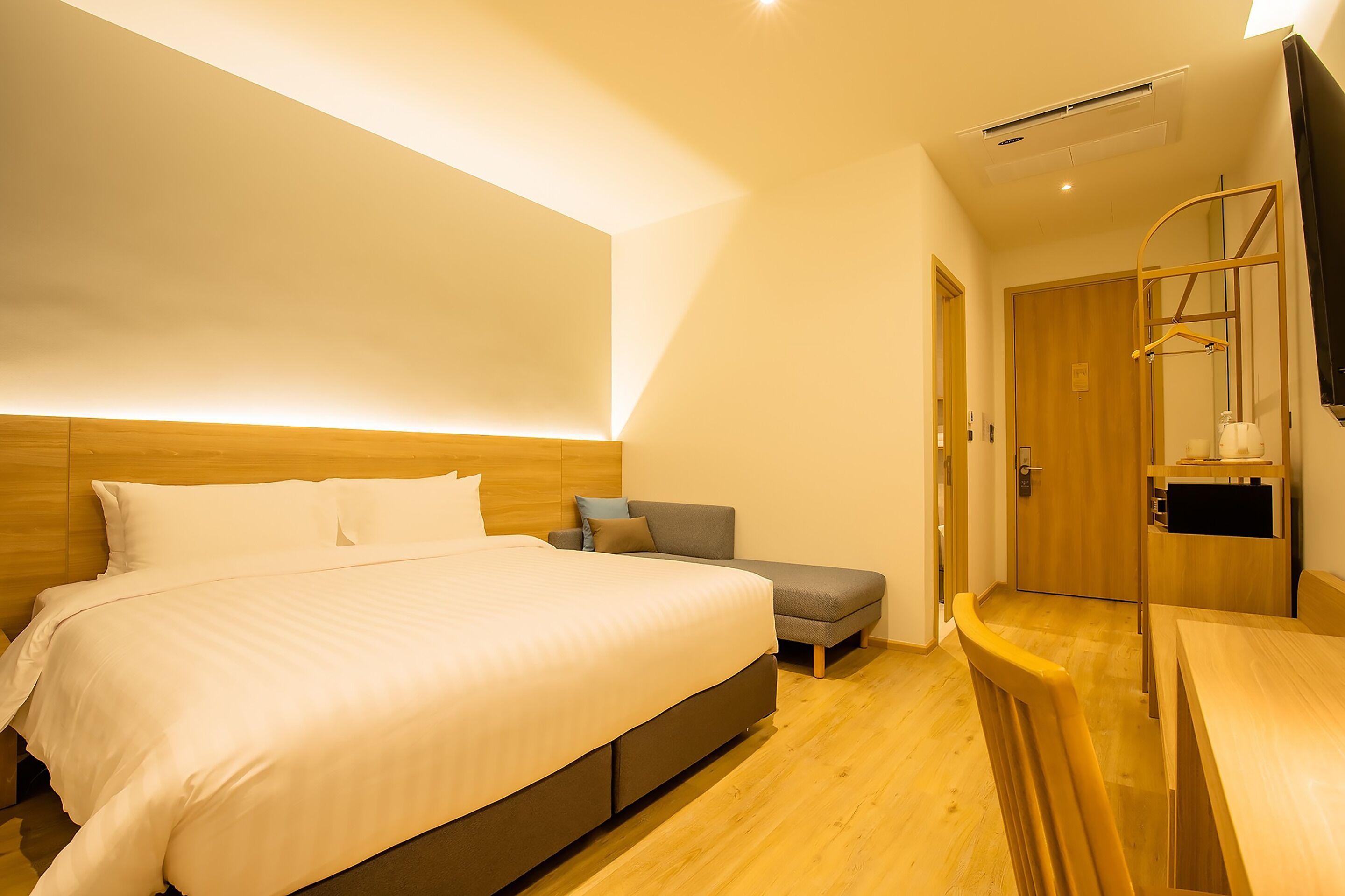 Un dormitorio moderno con una cama grande, cabecera de madera, un pequeño sofá, un escritorio de madera y un televisor montado en la pared.