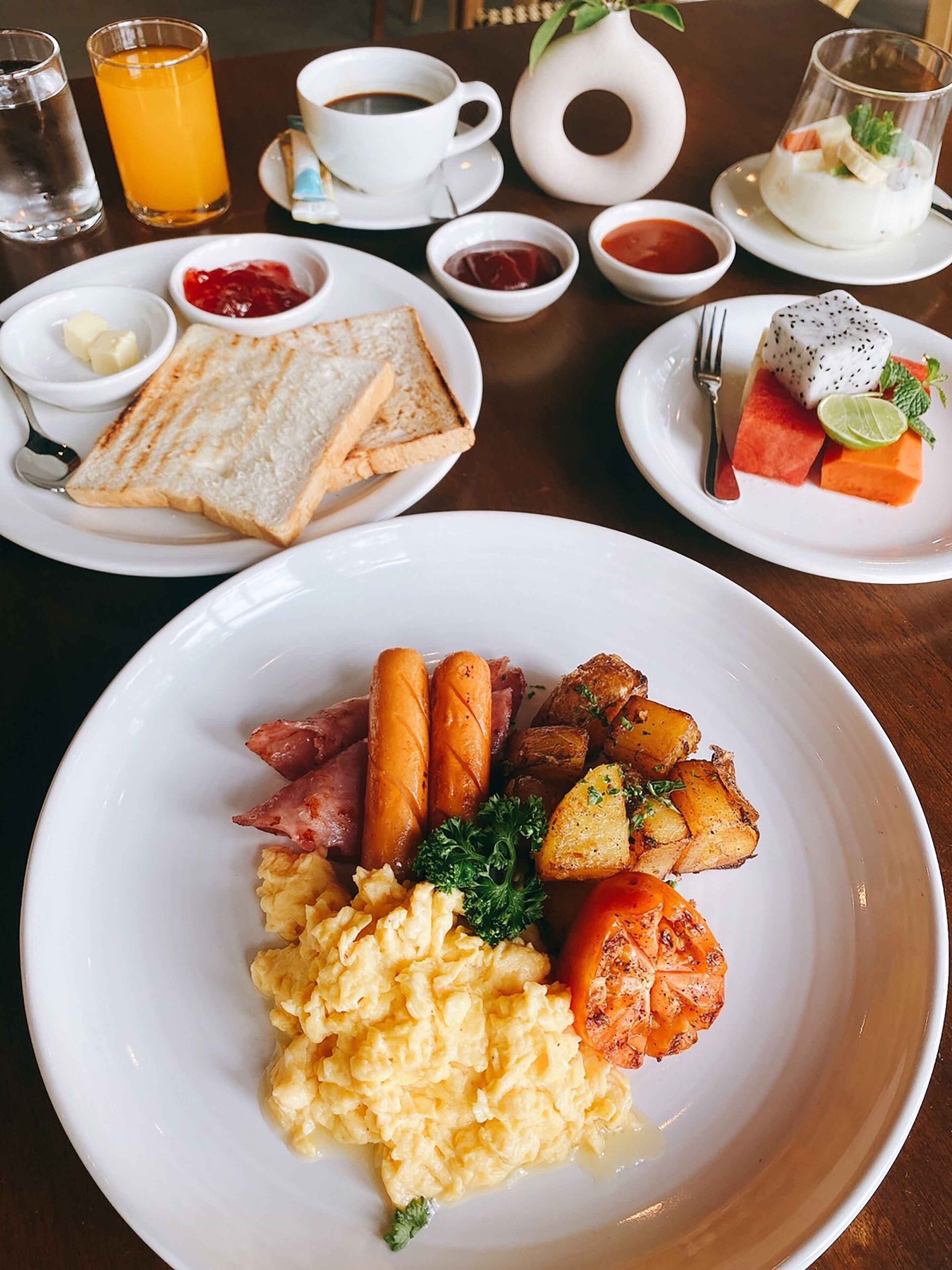 Un desayuno con tostadas, huevos revueltos, salchichas, papas asadas y una bandeja de fruta.
