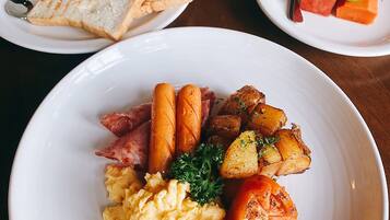 Un desayuno con tostadas, huevos revueltos, salchichas, papas asadas y una bandeja de fruta.