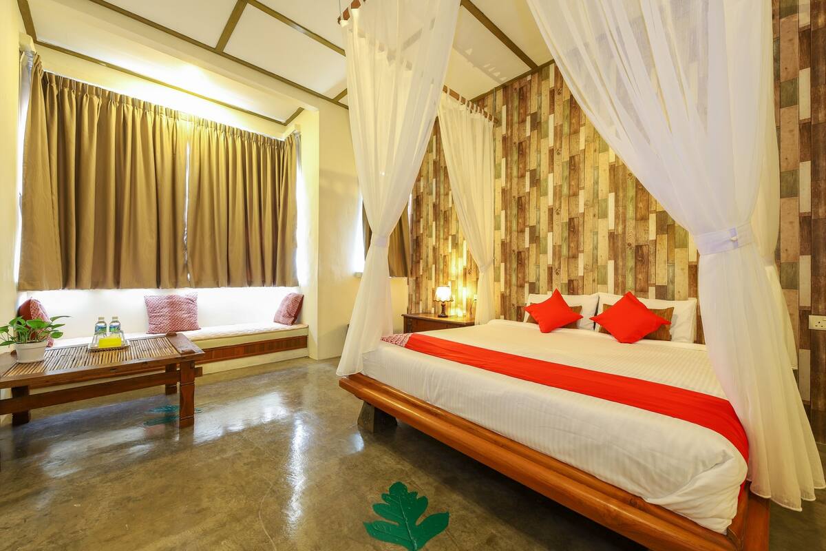A bedroom with a bed, a bedside table, a window with curtains, a wooden floor, and a wall with a patterned design.