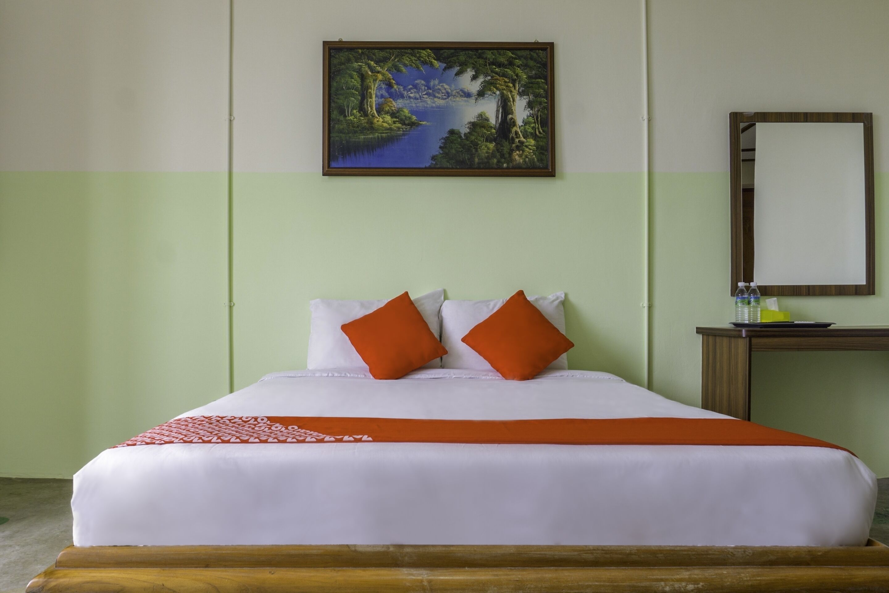 A neatly made bed with white linens and two orange pillows, a wooden headboard, a green wall, a painting of a landscape, and a small table with a mirror and a bottle of water.