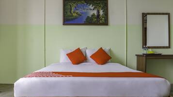 A neatly made bed with white linens and two orange pillows, a wooden headboard, a green wall, a painting of a landscape, and a small table with a mirror and a bottle of water.