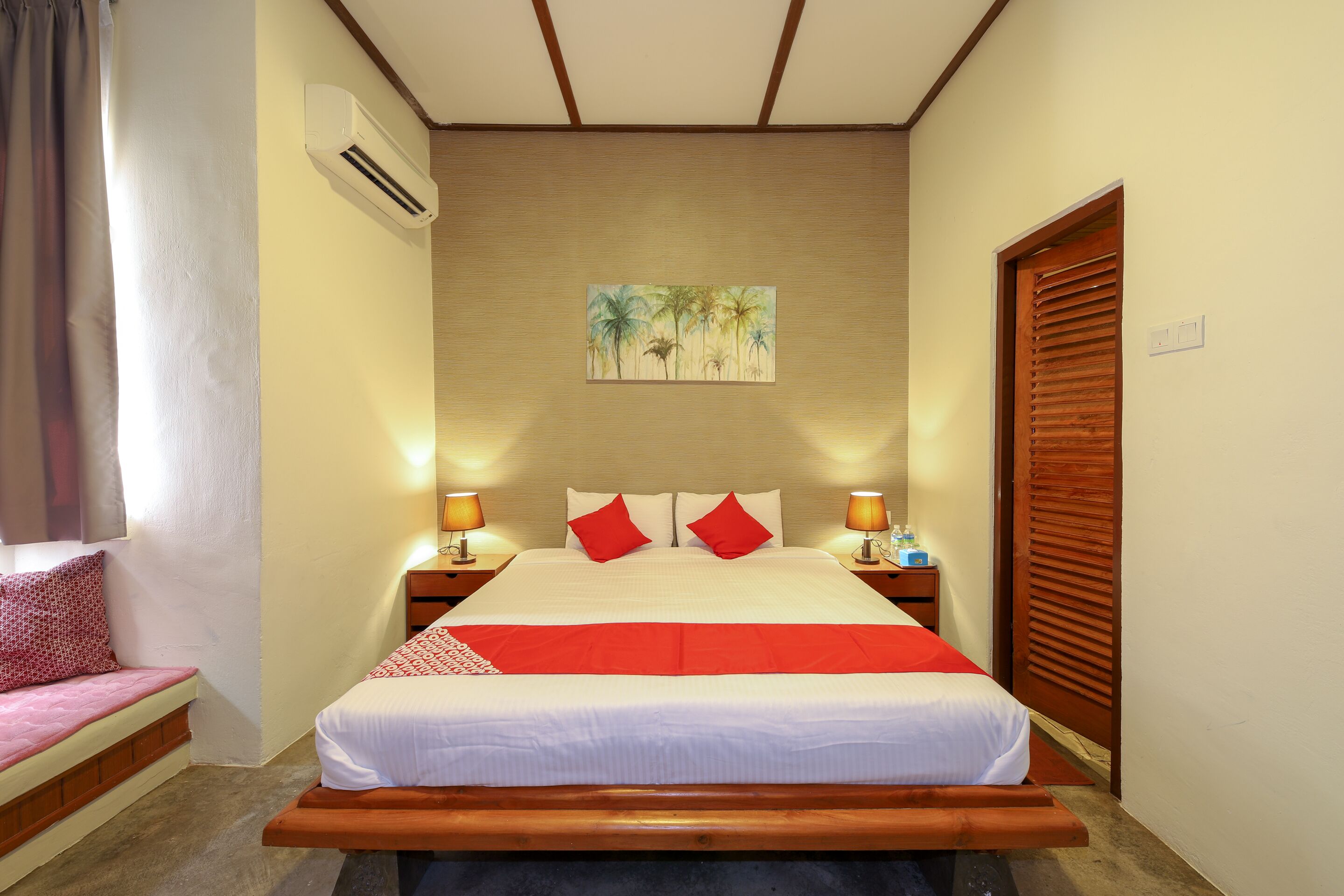 A hotel room with a large bed, a bench at the foot of the bed, a nightstand with a lamp, a wooden door, and a painting on the wall.