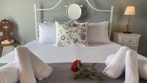 Een net opgemaakt bed met witte lakens, een bloemenkussen en een lamp in de vorm van een hartje op een nachtkastje.