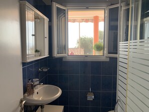 Un baño con un lavabo blanco, un espejo y una ventana que da a un edificio rojo y follaje exterior.