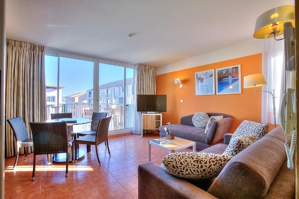A modern living room with a sofa, a coffee table, and a TV. There is a dining area with chairs and a table. The room has large windows and a view of buildings outside.
