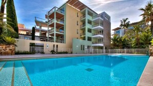 A swimming pool with a clear blue water, surrounded by a multi-story building with balconies, and palm trees in the background. - Apartment Port Prestige (Antibes)